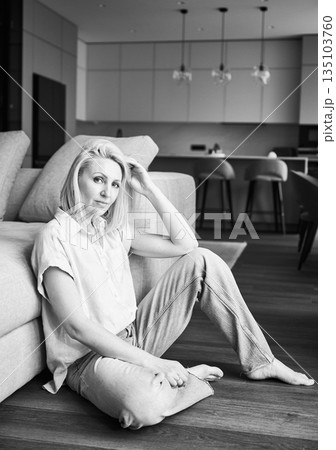 Portrait of woman sitting next to couch in her apartment. Pretty female in casual clothes looking straight on camera. Black and white image. Portrait of woman sitting next to couch in her apartment. Pretty female in casual clothes looking straight on camera. Black and white image. 135103760