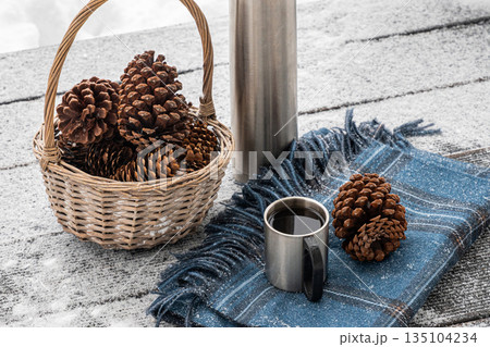 Thermos, metal mug, basket with pine cones and warm blanket on snowy wooden surface outdoors. Cozy winter picnic and outdoor relaxation concept. 135104234