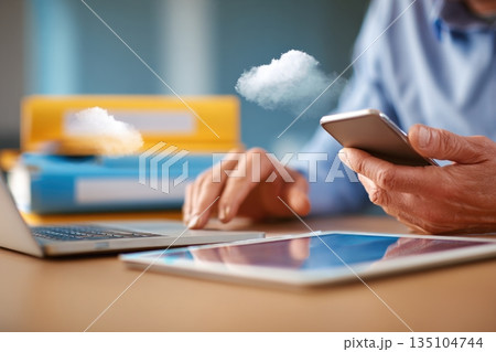 Hands using smartphone and tablet while working at a desk in a modern office with a laptop and cloud computing elements 135104744