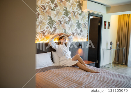 Woman in white bathrobe sitting on the bed in a hotel suit and looking relaxed Woman in white bathrobe sitting on the bed in a hotel suit and looking relaxed 135105070