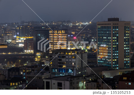 Night cityscape of Pristina, capital of Kosovo, showing modern high rise buildings, residential neighborhoods, illuminated windows, urban density and foggy winter atmosphere 135105253