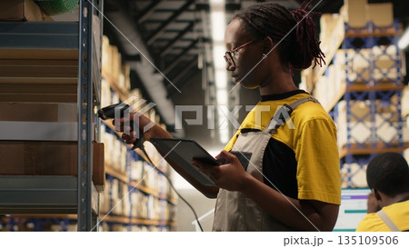 Black woman staff using a barcode scanner to process shipping labels on boxes from storage racks. Scanning awb tags for package tracking service with tracking info, accurate logistics. Camera B. 135109506