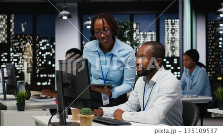 Black company advisors doing teamwork at night to meet deadlines, completing monthly reports and KPI numbers for business productivity. Tasks in strategy room after hours. 135109537