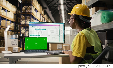 Female warehouse clerk checking awb labels next to chroma key on screen, working in depot with industrial storage racks. Fulfillment center deal with distribution and e-commerce. Camera A. 135109547