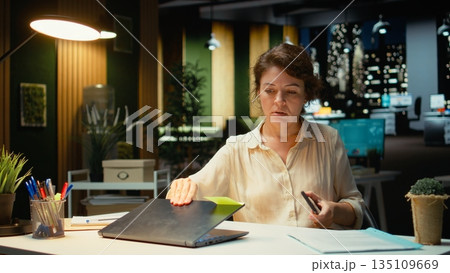Female employee checking the time on her phone and closing laptop, preparing to leave office late at night after doing overtime. Intense workload makes her stay after hours. Camera A. 135109669