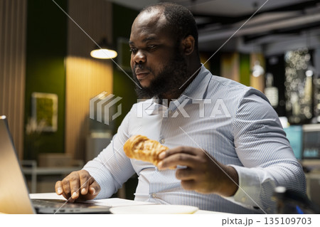 Black man analyst does emotional compulsive overeating due to workload at workstation, eating junk food and pastry as an unhealthy lifestyle. Emotional stress eating disorder at night. Black man analyst does emotional compulsive overeating due to workload at workstation, eating junk food and pastry as an unhealthy lifestyle. Emotional stress eating disorder at night. 135109703