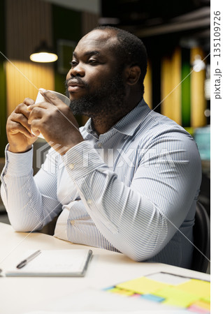 African american guy drinking coffee late at night to finish his work, sitting at desk alone at the workspace. Businessman meeting deadlines with tasks and reports, management. 135109726