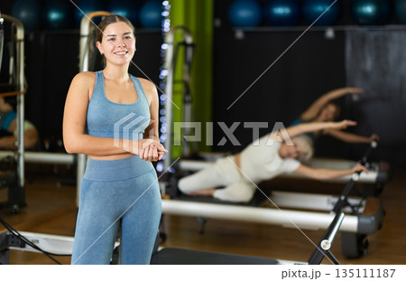 Female trainer stands in gym against background of Pilates reformer machines Female trainer stands in gym against background of Pilates reformer machines 135111187