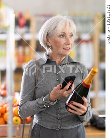 Checking expiration date of sparkling wine - elderly female shopper scans QR code on label of wine using smartphone 135111221