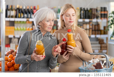 Girl and grandmother buyers examine good, doubt and choose between varieties of fruit juice in store 135111405
