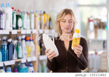 Girl housewife in household store examines goods, chooses hair care product Girl housewife in household store examines goods, chooses hair care product 135111601