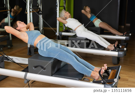 Young woman sitting on box set equipment reformer performs for abdominal muscles Young woman sitting on box set equipment reformer performs for abdominal muscles 135111656