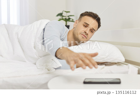 Young sleepy man waking up in the morning and stretching to mobile phone lying in bed at home. 135116112