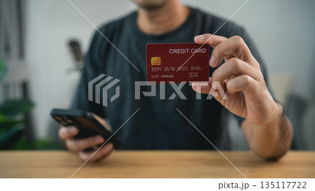 Man showing red credit card to camera while holding smartphone. Concept of financial security, banking identity verification, credit score, e-commerce payment, and digital economy. 135117722