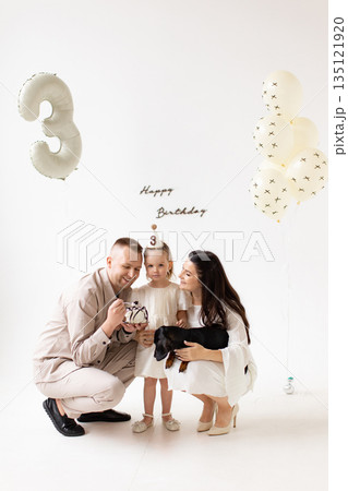 A family celebrates a little girl's third birthday with a cake and a dachshund dog. A family celebrates a little girl's third birthday with a cake and a dachshund dog. 135121920