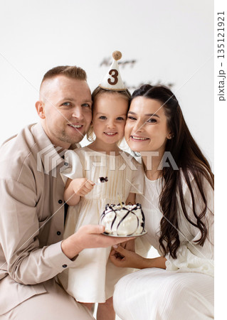 A happy family of three celebrates a little girl's third birthday with a cake and party hat. A happy family of three celebrates a little girl's third birthday with a cake and party hat. 135121921