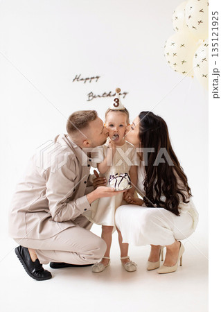 A young girl celebrates her third birthday with her parents, enjoying cake and kisses in a bright, cheerful setting. A young girl celebrates her third birthday with her parents, enjoying cake and kisses in a bright, cheerful setting. 135121925