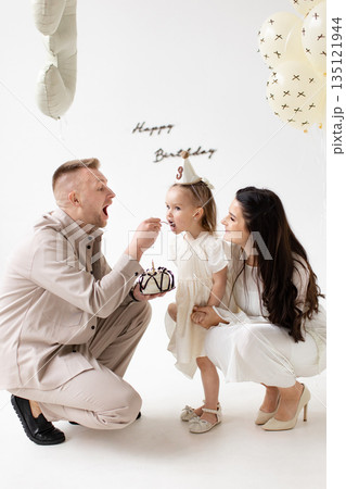 A family of three celebrates a little girl's birthday with cake and balloons in a bright, white room. A family of three celebrates a little girl's birthday with cake and balloons in a bright, white room. 135121944