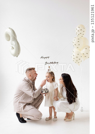 A family celebrates a little girl's birthday with cake and balloons in a bright, white room. A family celebrates a little girl's birthday with cake and balloons in a bright, white room. 135121961
