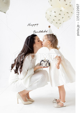 A tender moment captured as a mother kisses her daughter during a birthday celebration, complete with a cake and balloons. 135122007