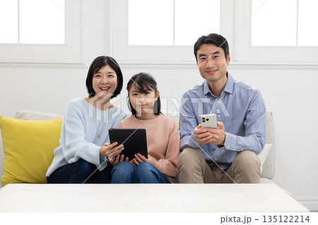 ソファに座り笑顔でタブレットとスマートフォンを操作する3人家族 135122214