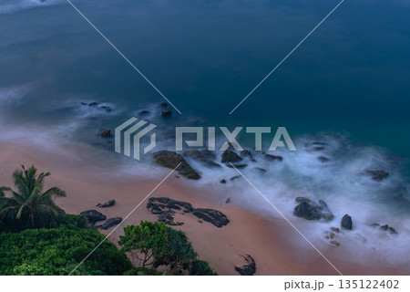 A birds eye view of a beach in Sri Lanka, featuring rocks scattered along the shoreline and water crashing against the rocks. A birds eye view of a beach in Sri Lanka, featuring rocks scattered along the shoreline and water crashing against the rocks. 135122402