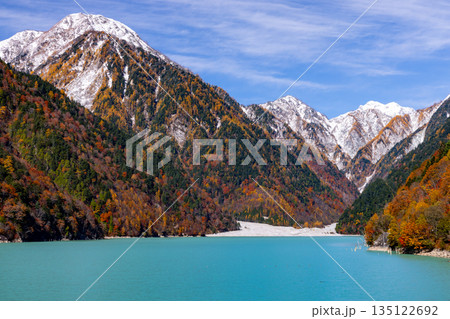 長野県大町市平　鮮やかなミルキーブルーの高瀬ダム調整湖の紅葉と青空に映える冠雪した山の景色 135122692
