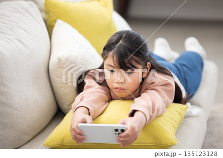 ソファに寝転んでスマートフォンを操作する小学生の女の子 135123128