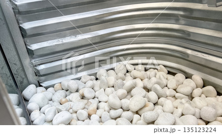 Corner section of a corrugated metal window well filled with small white decorative rocks. The texture of the galvanized steel contrasts with the smoothness of the pebbles under natural daylight. 135123224