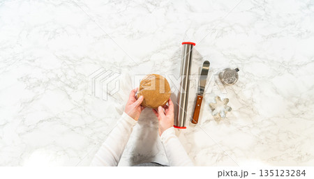 Overhead view of Amber Gingerbread Cookies dough, a rolling pin, cookie cutters, and flour shaker placed neatly on a marble surface, ready for preparation. 135123284
