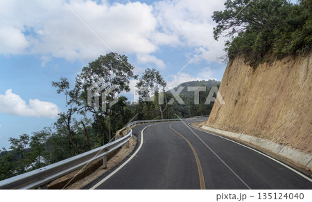 Scenic winding asphalt road on a lush green mountain under a beautiful blue sky with clouds. Perfect for travel, road trip, journey, and transportation infrastructure concept 135124040