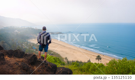 Man with backpack stands on a rock looking at the sea Goa India Man with backpack stands on a rock looking at the sea Goa India 135125110