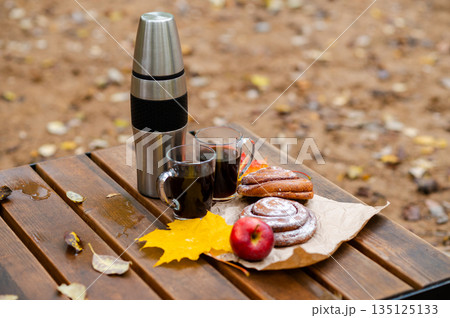 Autumn picnic setup with thermos, coffee mugs, cinnamon rolls, apple, and yellow maple leaves on a wooden bench outdoors Autumn picnic setup with thermos, coffee mugs, cinnamon rolls, apple, and yellow maple leaves on a wooden bench outdoors 135125133