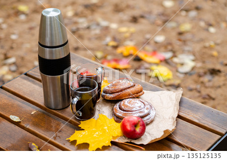 Autumn picnic setup with thermos, coffee mugs, cinnamon rolls, apple, and yellow maple leaves on a wooden bench outdoors Autumn picnic setup with thermos, coffee mugs, cinnamon rolls, apple, and yellow maple leaves on a wooden bench outdoors 135125135