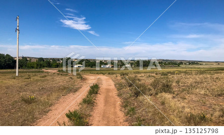 Dusty dirt road leading through a peaceful rural village with scattered houses and lush green trees under a clear blue sky. Dusty dirt road leading through a peaceful rural village with scattered houses and lush green trees under a clear blue sky. 135125798