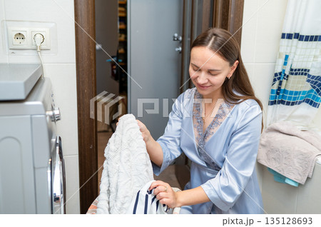 Beautiful housewife in silk robe sorting dirty laundry by washing machine at home 135128693