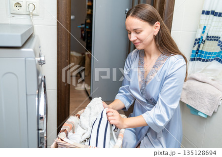 Beautiful housewife in silk robe sorting dirty laundry by washing machine at home 135128694