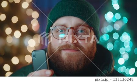 Young man in beanie holds smartphone, smiles against festive holiday lights background 135130249