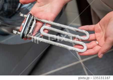 Hands holding washing machine heating element covered with limescale during appliance repair 135131187
