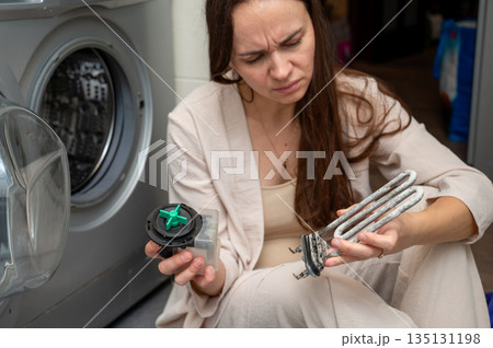 Woman examining washing machine parts  heating element and pump motor during home appliance repair 135131198