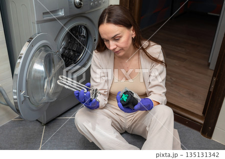 Woman examining washing machine parts  heating element and pump motor during home appliance repair 135131342