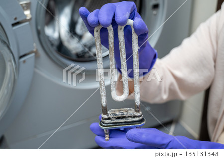 Person in gloves showing removed washing machine heating element covered with limescale 135131348