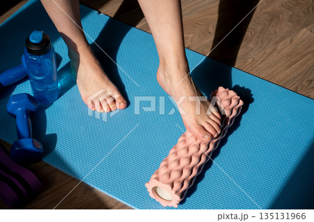 Female legs on foam roller doing myofascial massage with dumbbells and water bottle nearby 135131966