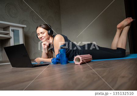 Woman lying on yoga mat with laptop headphones dumbbells water bottle and foam roller at home 135132510