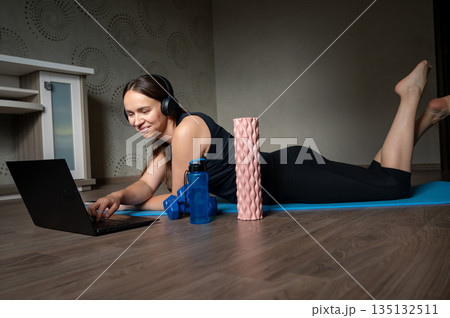 Happy woman lying on yoga mat with laptop headphones dumbbells water bottle and foam roller at home 135132511