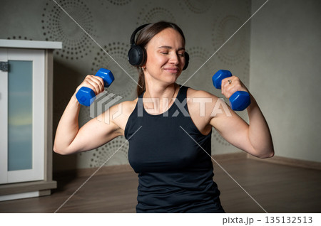Smiling woman training with dumbbells at home wearing headphones 135132513