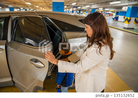 Mother opening rear car door and helping child get into vehicle in public parking garage. Family travel routine, childcare support and everyday mobility moment in real life urban environment Mother opening rear car door and helping child get into vehicle in public parking garage. Family travel routine, childcare support and everyday mobility moment in real life urban environment 135132652