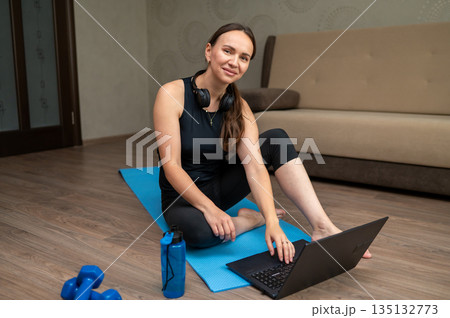 Woman sitting on yoga mat at home watching laptop before fitness workout Woman sitting on yoga mat at home watching laptop before fitness workout 135132773