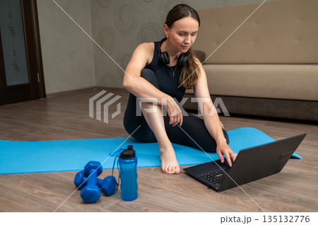 Woman sitting on yoga mat at home watching laptop before online pilates workout Woman sitting on yoga mat at home watching laptop before online pilates workout 135132776
