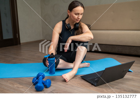 Woman sitting on yoga mat at home watching laptop before online pilates workout 135132777